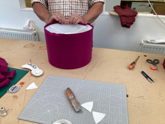 A close-up of a participant's hands carefully wrapping fabric around a lampshade frame during a lampshade-making workshop. Various fabric swatches, scissors, and other crafting tools are visible on the table, showcasing the hands-on learning process.