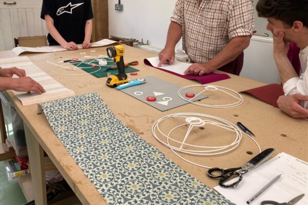 A group of students working on their their newly crafted lampshades, each with a unique design during a lampshade-making course. The workshop setting includes tables covered with fabrics and tools, capturing the creative atmosphere of the class.