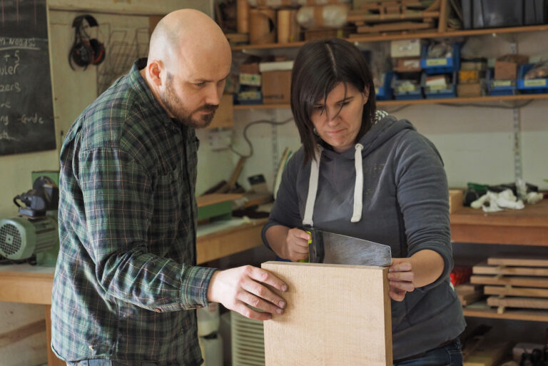 Teaching a student woodworking. The teacher is holding some wood and the student is cutting.